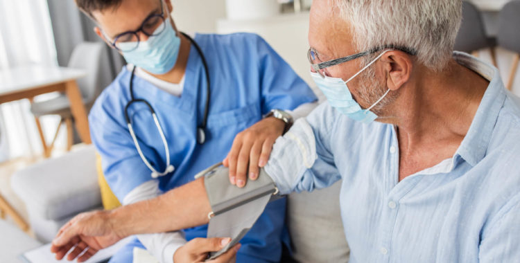 Male nurse measures blood pressure to senior man with mask while being in a home visit.