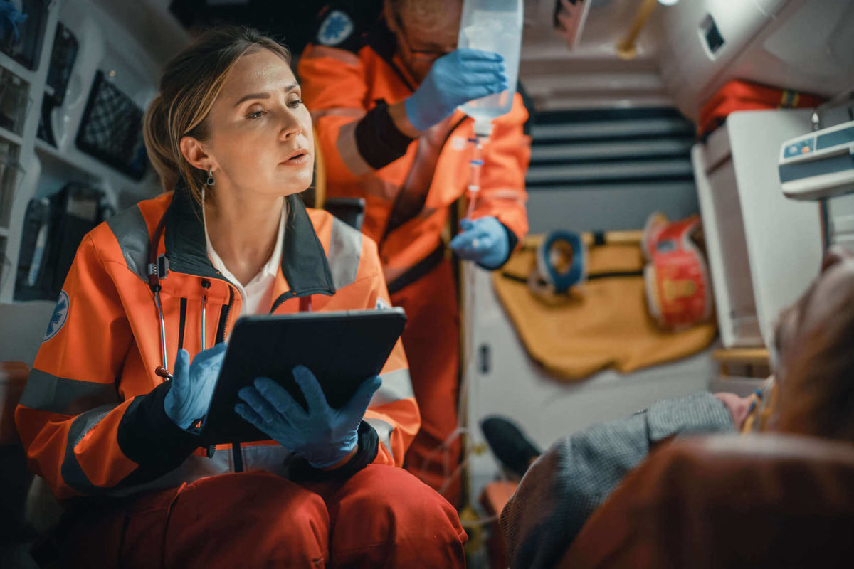 Female EMS Professional Paramedic Using Tablet Computer to Fill a Questionnaire for the Injured Patient on the Way to Hospital. Emergency Care Assistant Comforting the Patient in an Ambulance.