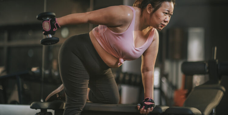 body positive Asian mid adult woman exercising with dumbbells in a lunge position at gym bench at night