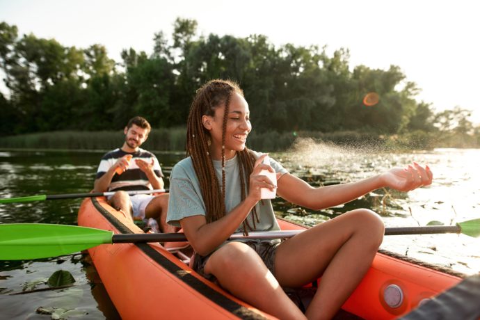 Cheerful young woman spraying sunscreen protection while kayaking on the river with her boyfriend outdoors