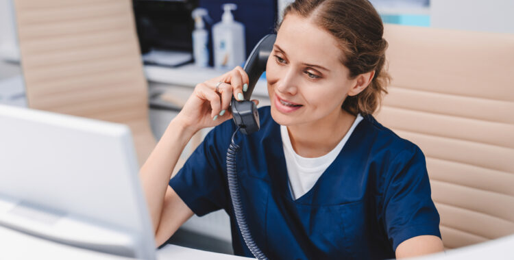 Young female receptionist talking on phone in clinic while sitting and looking on pc monitor