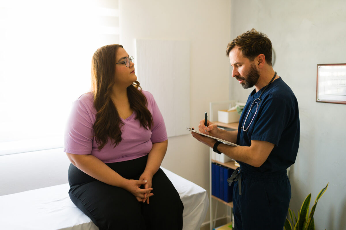 Plus-size female patient conversing with healthcare professional, taking notes during medical consultation in clinical setting