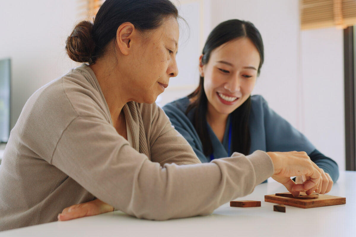 Asian caregiver nurse wearing blue scrubs and elderly woman retirement playing wooden shape puzzles game learning origami as for physiotherapy activity for dementia as a part of a physical therapy session Medical health care plan