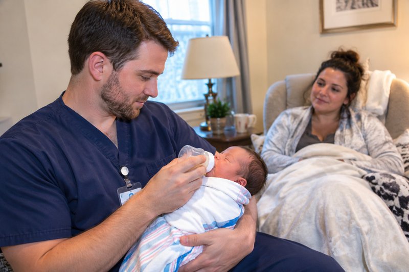 Male nurse feeding newborn while the mother rests on a chair